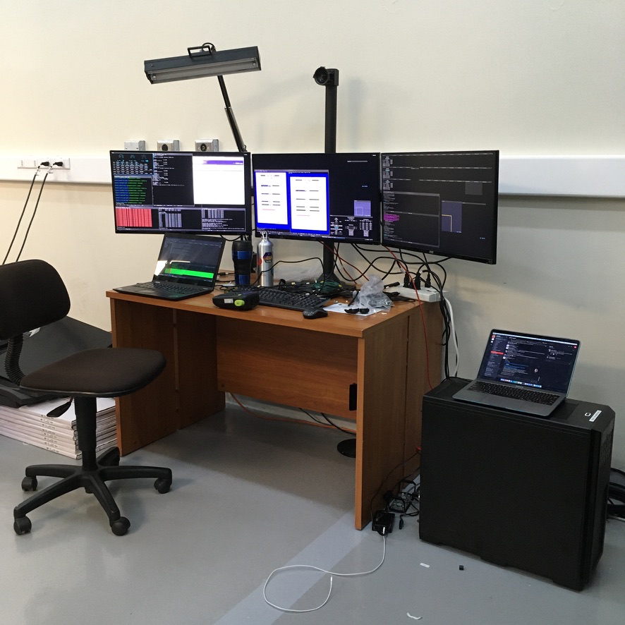 The MagAO-X Megadesk with three monitors dominates a work table. A lonely laptop is balanced atop a workstation (right) as a sort of makeshift desk.