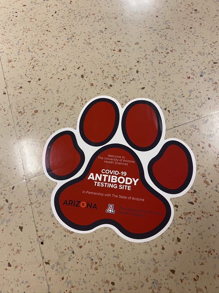 A large paw print sticker modeled on a wildcat, affixed to a tile floor. It reads "Welcome to The University of Arizona Health Sciences COVID-19 Antibody Testing Site In Partnership with The State of Arizona." with logos for both Arizona and the University.