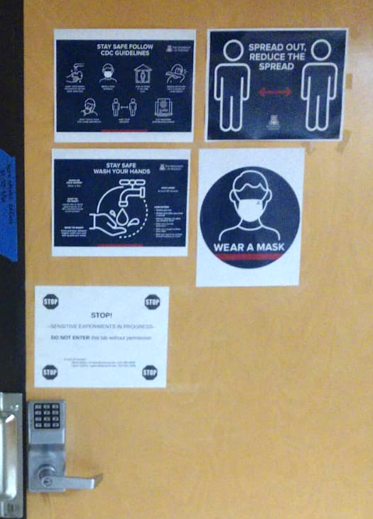Picture of the door to the lab with four navy blue signs reiterating CDC guidelines for infection control and one sign saying "do not enter"