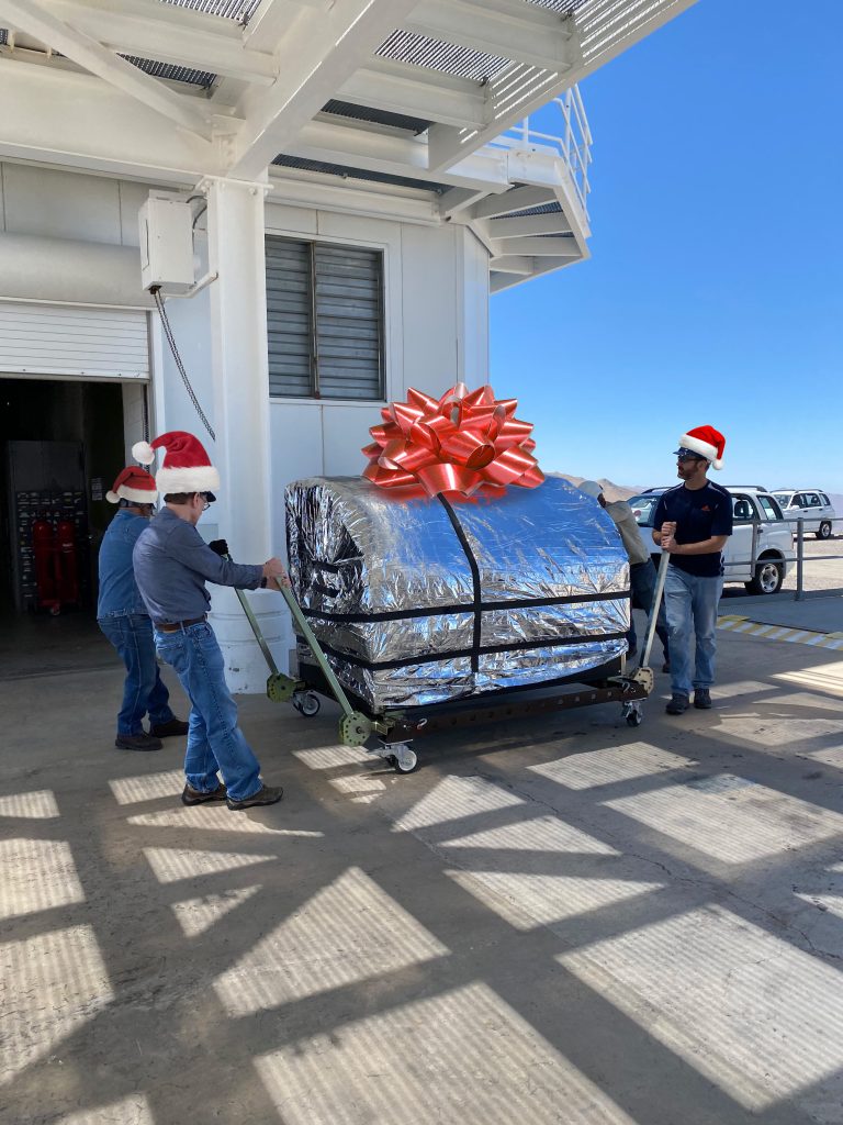 Jared, Laird, and a Las Campanas engineer move the foil-wrapped MagAO-X instrument on a cart. They're wearing photoshopped Santa Claus hats, and the MagAO-X instrument has a photoshopped bow on it.